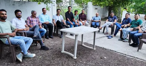 A group of men sit in a circle of plastic chairs around a white table at an outdoor gathering.
