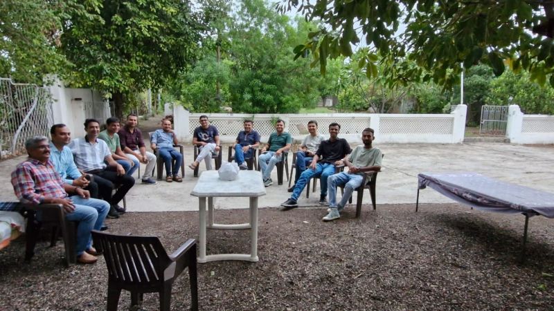 A group of men sit in a circle on plastic chairs under trees in an open courtyard.