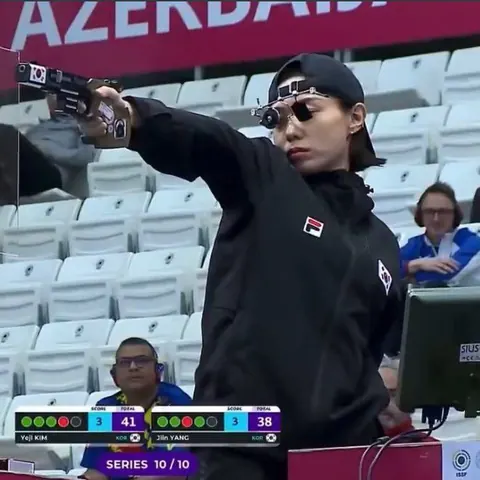 A woman in a black cap and jacket aims a pistol with specialised shooting glasses at an indoor range, scoreboard below.
