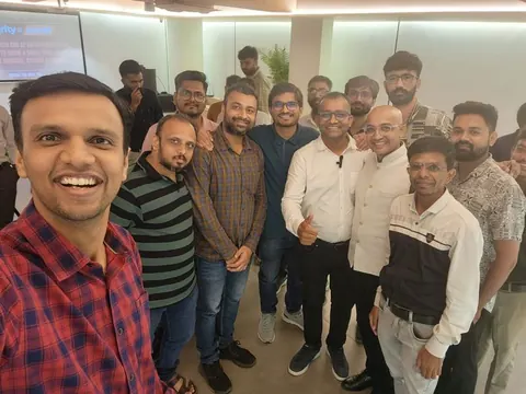 Group selfie of smiling men at an indoor tech meetup with a presentation screen in the background.