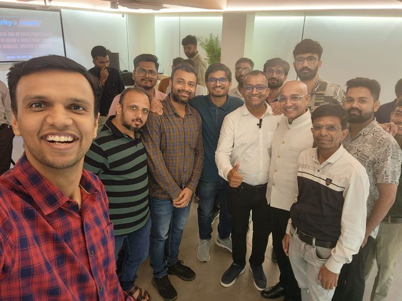 Group selfie of smiling men at an indoor tech meetup with a presentation screen in the background.
