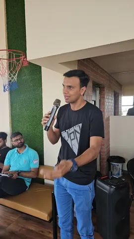 A man in a black T-shirt speaks into a microphone at an indoor event, two attendees seated on a bench behind him.
