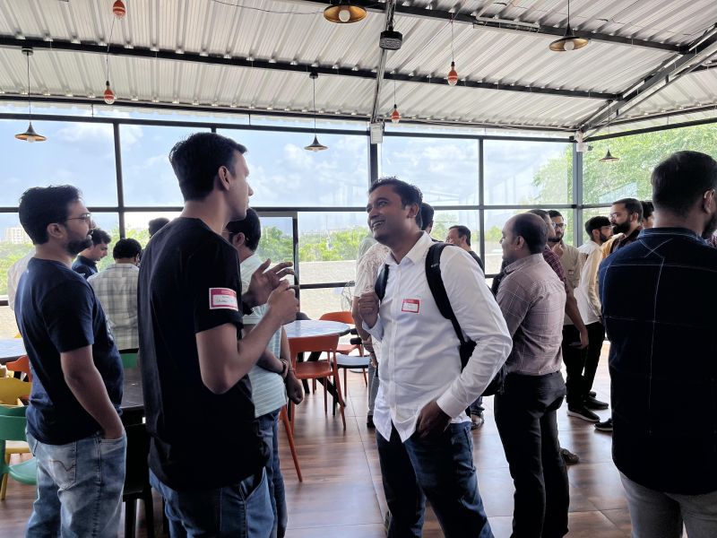 Men wearing name badges chat in small groups on a covered terrace with glass walls at a tech meetup.