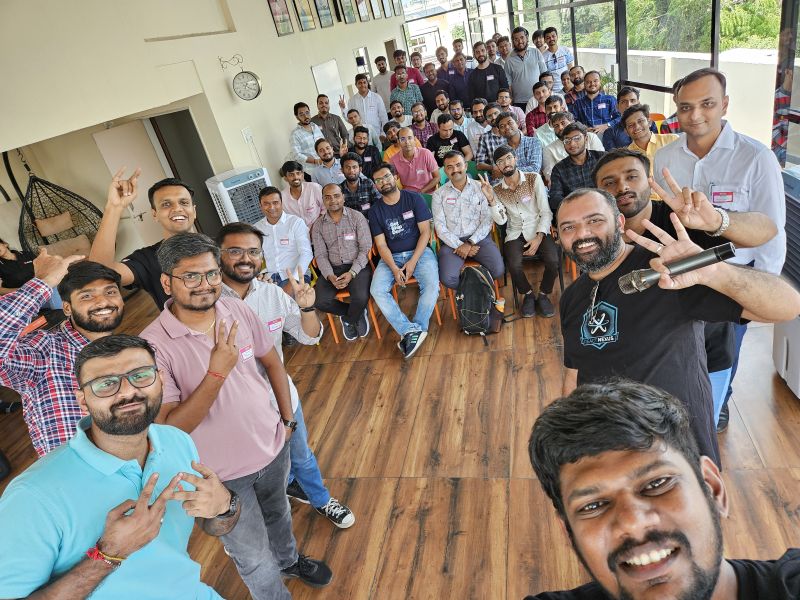 A large group of men pose for a selfie at an indoor tech event, many holding up peace signs.