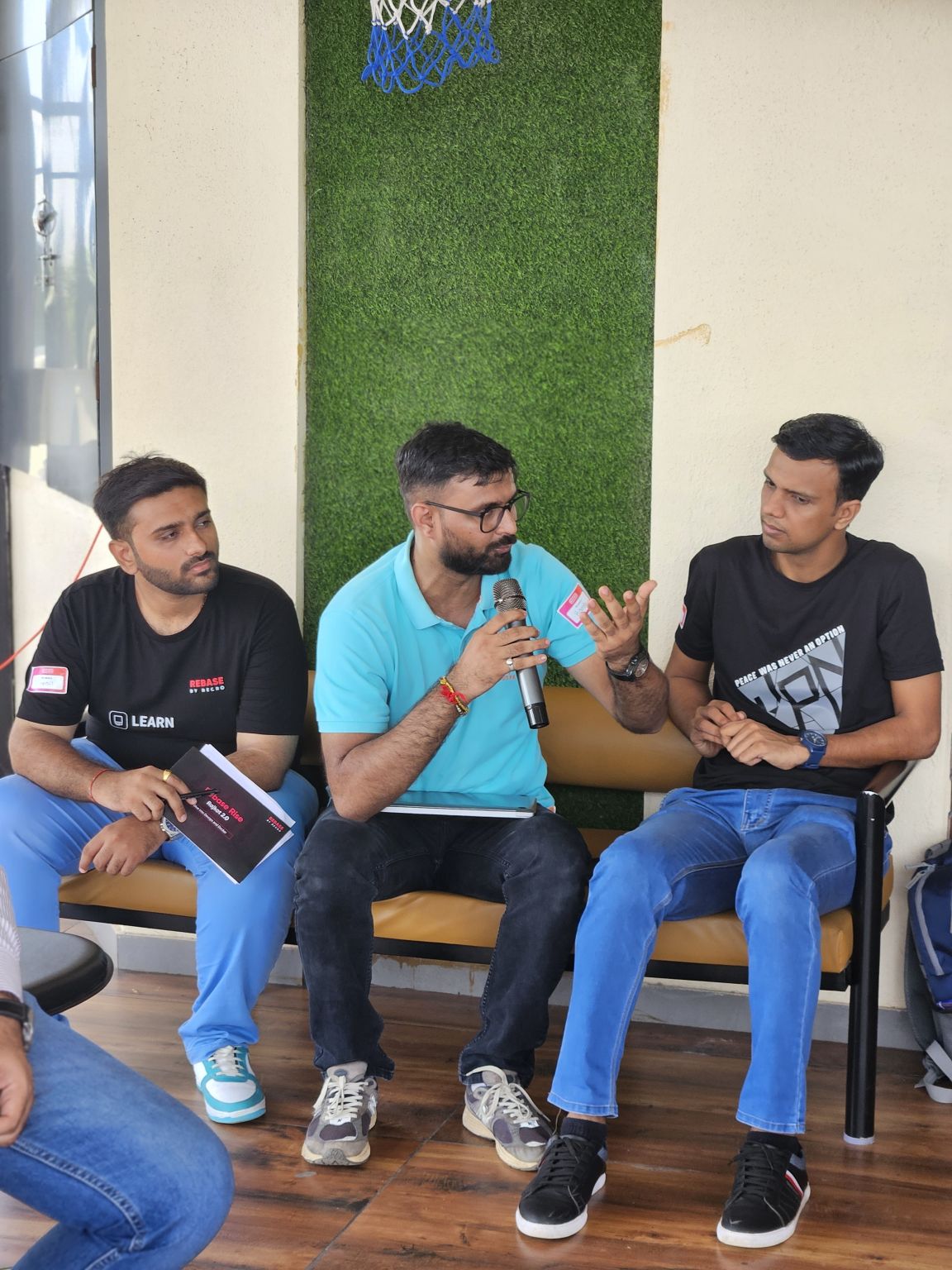 A man in a turquoise polo speaks into a microphone on a bench, flanked by two seated men at a meetup.