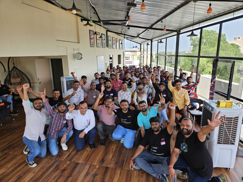 A large group of men cheer and pose together for a group photo on a covered terrace at a community event.