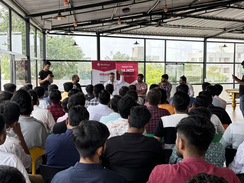 A panel of speakers faces a seated audience at a tech event, with red banners on the wall behind them.