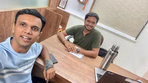 A man in a blue t-shirt takes a selfie with another man seated smiling at an office desk with a flask and laptop.