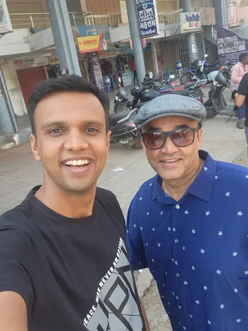 Selfie of two smiling men on a street with parked scooters and shops with Gujarati signage in the background.