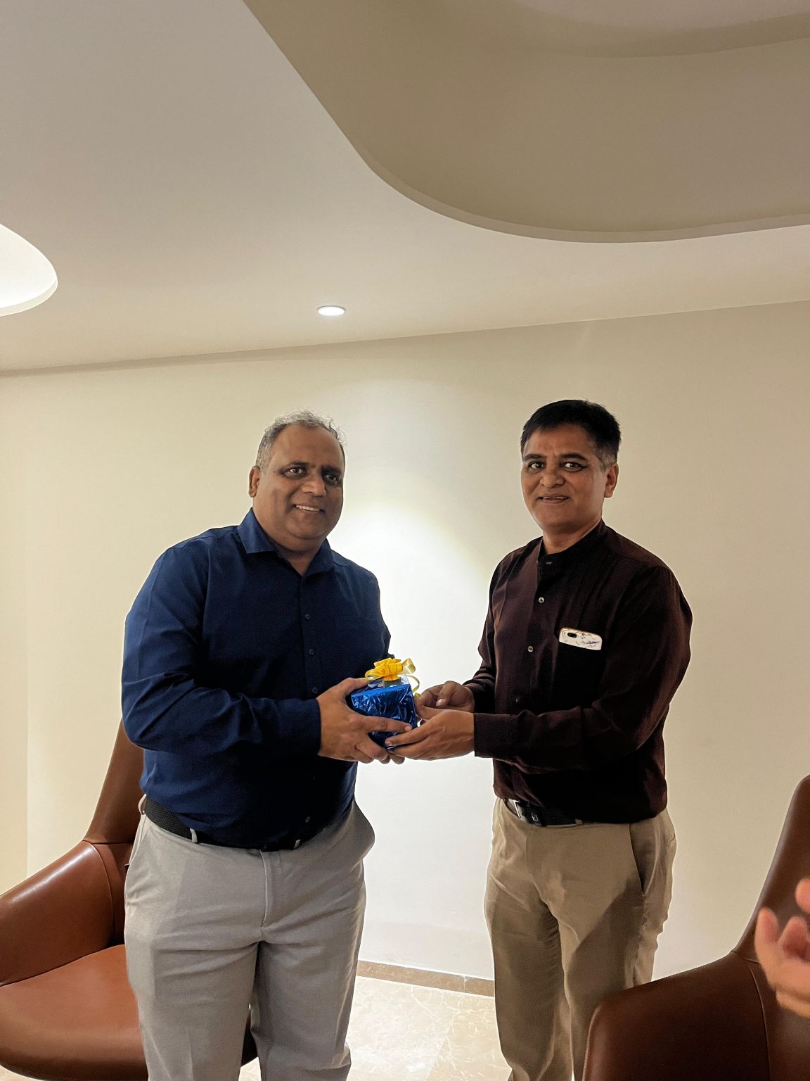 Two men smiling while one hands over a small blue gift box tied with a yellow ribbon