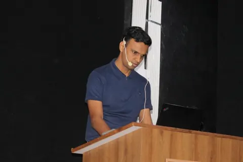 Gaurav speaking at a wooden lectern with a laptop, wearing a headset microphone against a black backdrop.