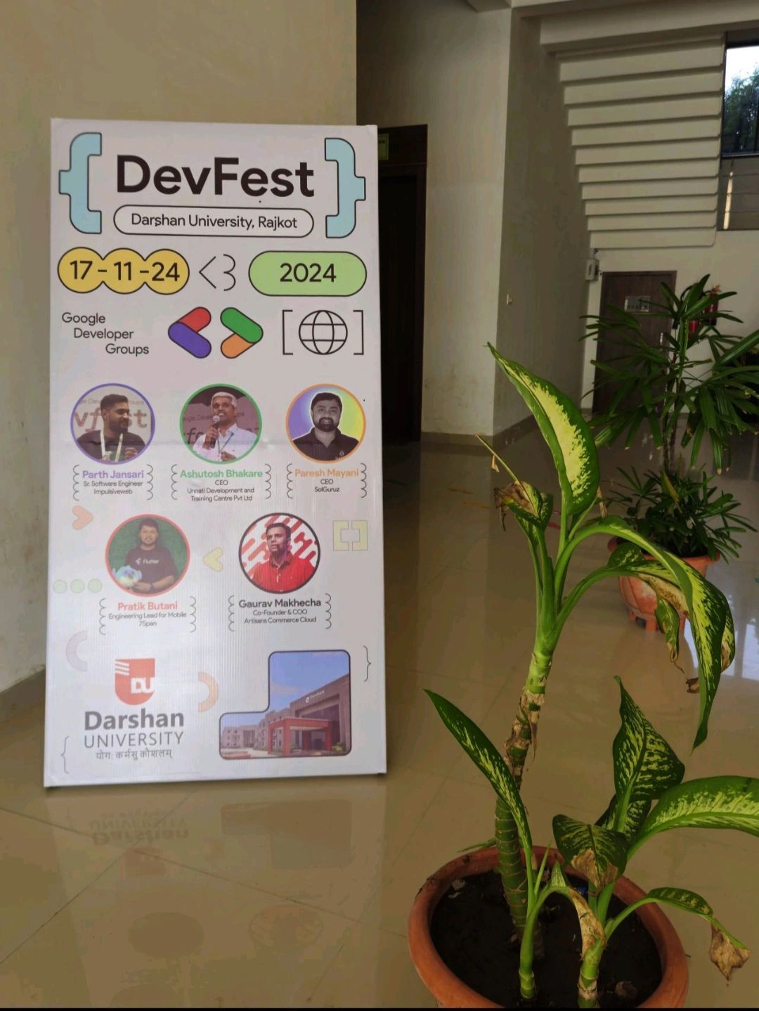 DevFest banner at Darshan University, Rajkot profiling the event speakers, beside a potted plant.