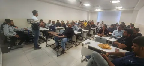 A man gestures while speaking to a seated audience in a classroom-style room at a community meetup.