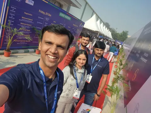 A man takes a selfie with three companions on a red carpet beside an event agenda board at a tech expo.
