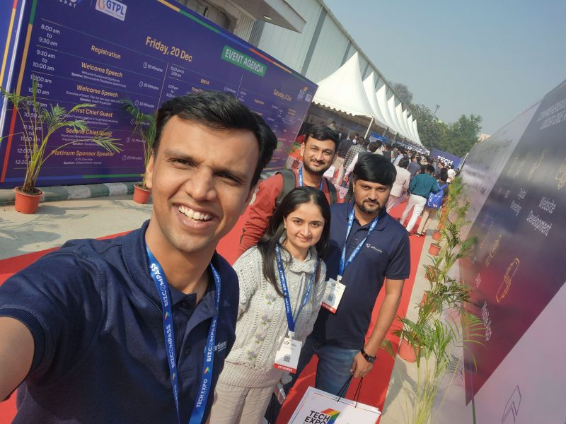A man takes a selfie with three companions on a red carpet beside an event agenda board at a tech expo.