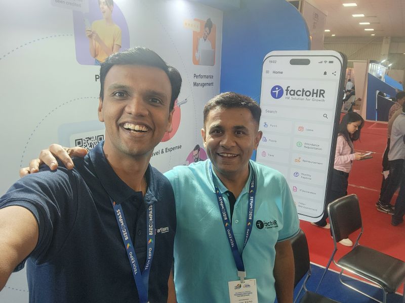A man takes a selfie with another man in a factoHR shirt at the factoHR booth at a tech expo.