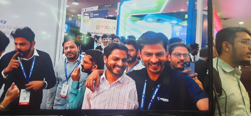 A screen at the expo shows a group of smiling men wearing Tech Expo lanyards in a crowded exhibition hall.