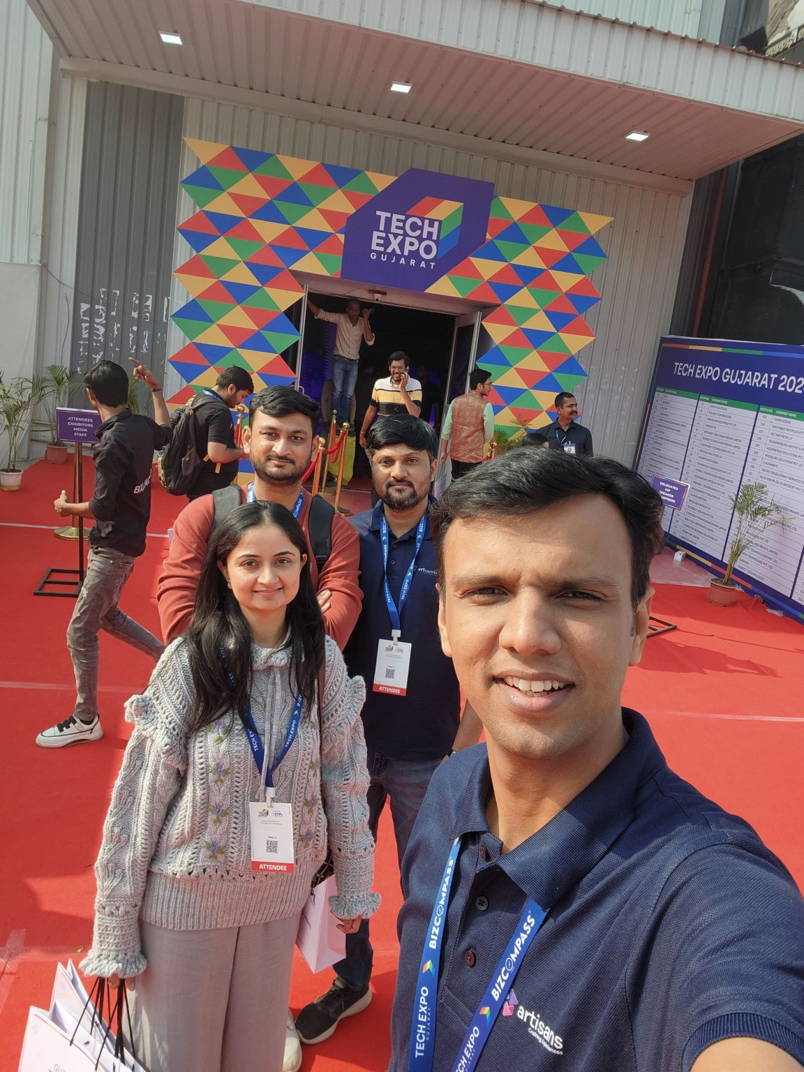 A man takes a selfie with three friends at the colourful Tech Expo Gujarat entrance on a red carpet.