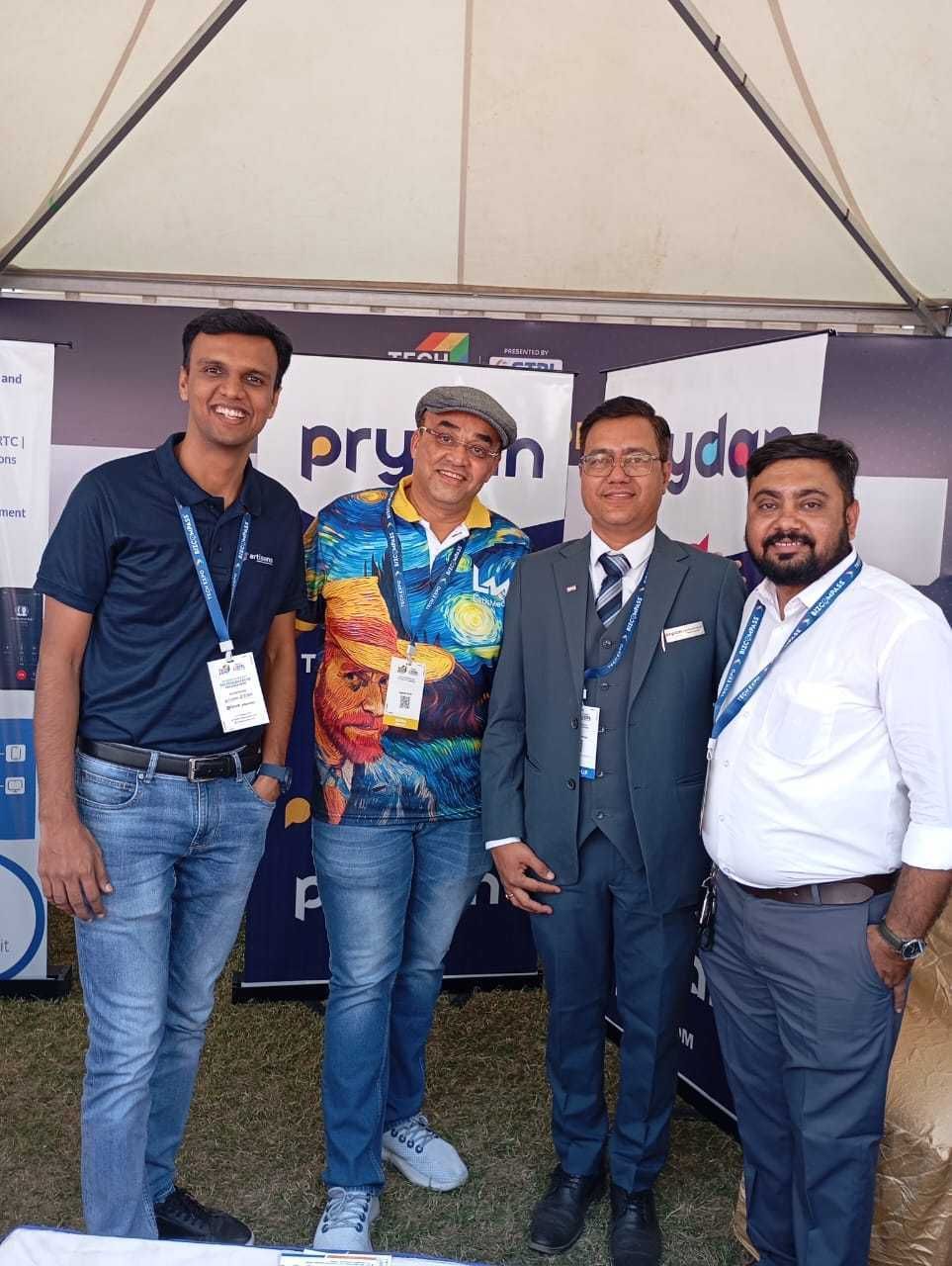 Four men wearing event lanyards pose together under a tent in front of Prydan roll-up banners.