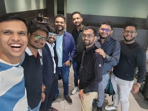 Eight men smile and pose together for a group selfie at an indoor event.