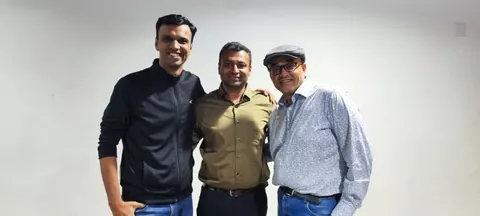 Three men stand side by side smiling against a plain white wall, one wearing a flat cap and patterned shirt.
