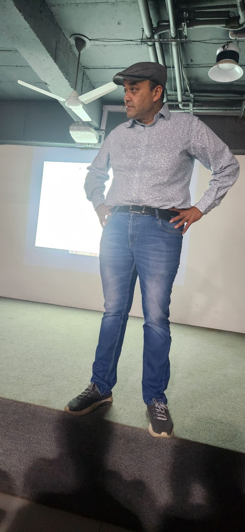 Man in a flat cap stands with hands on hips in front of a projection screen in a seminar room.