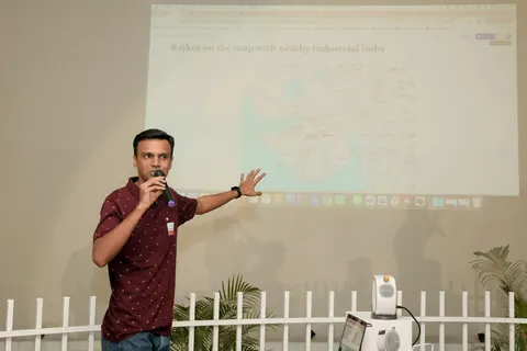 A man with a microphone gestures toward a projected map titled Rajkot on the map with nearby industrial hubs.