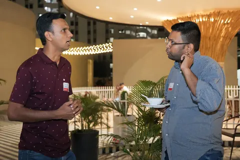 Two men wearing name badges talk at an evening event while one holds a plate with a bowl.