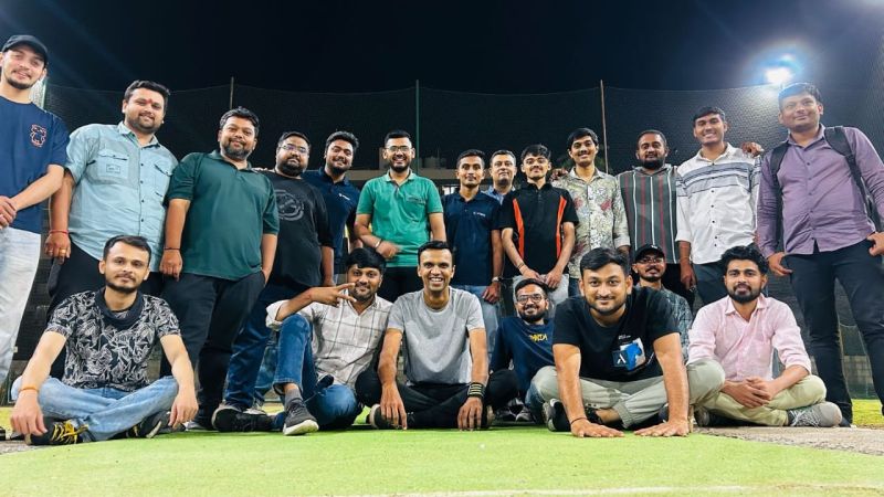 Large group of men posing together on a floodlit green turf at night, some seated in the front rows.