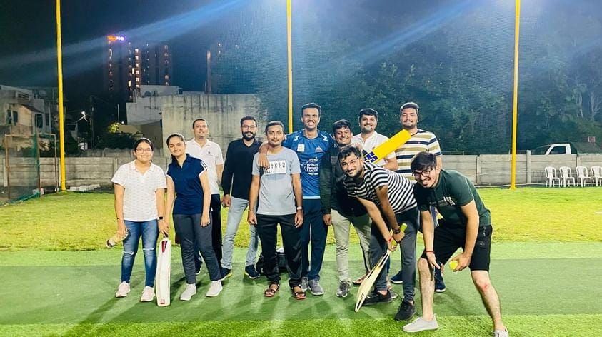 Small group of men and two women holding cricket bats on a floodlit green turf at night.