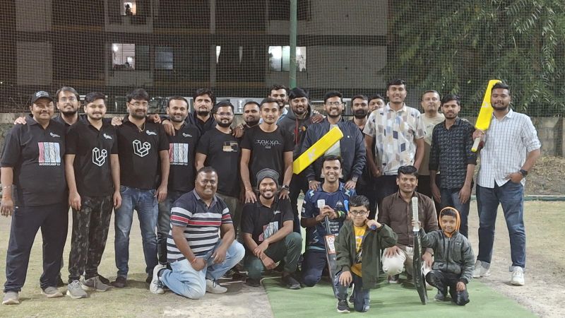 Big group of men and two children on turf at night, several in matching black t-shirts with yellow cricket bats.