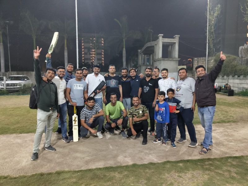 Group of men and boys posing at night on an open ground, one raising a cricket bat, another giving a thumbs up.