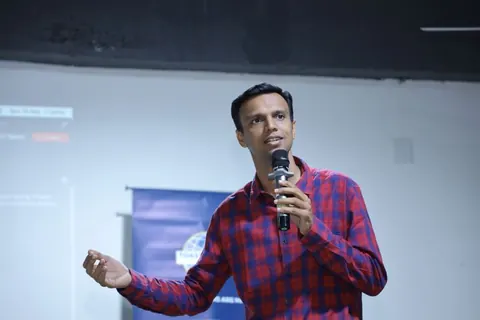 A man in a red and blue checked shirt speaks into a microphone on a stage with a banner and projected slide behind him.