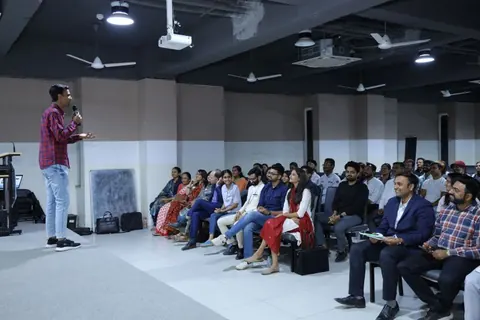 A man with a microphone addresses rows of seated attendees in a seminar hall with ceiling fans and a projector.