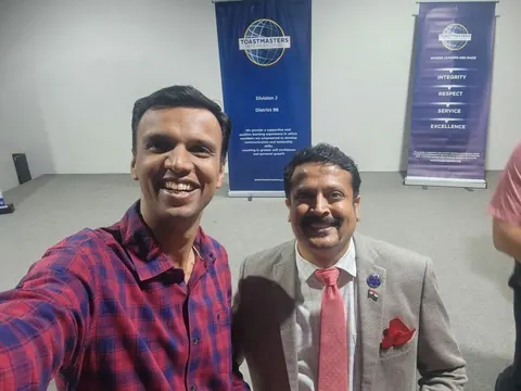 A smiling man takes a selfie with a man in a grey blazer and red tie in front of Toastmasters banners.