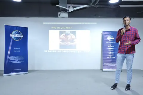 A man presents with a microphone beside a projected slide reading 'Why are you there?' between two Toastmasters banners.