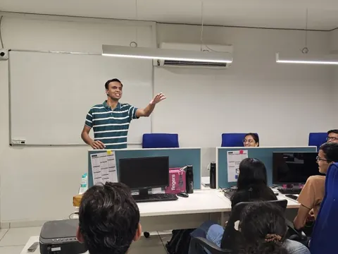 A man in a striped polo gestures while speaking to colleagues seated at office desks with computers.
