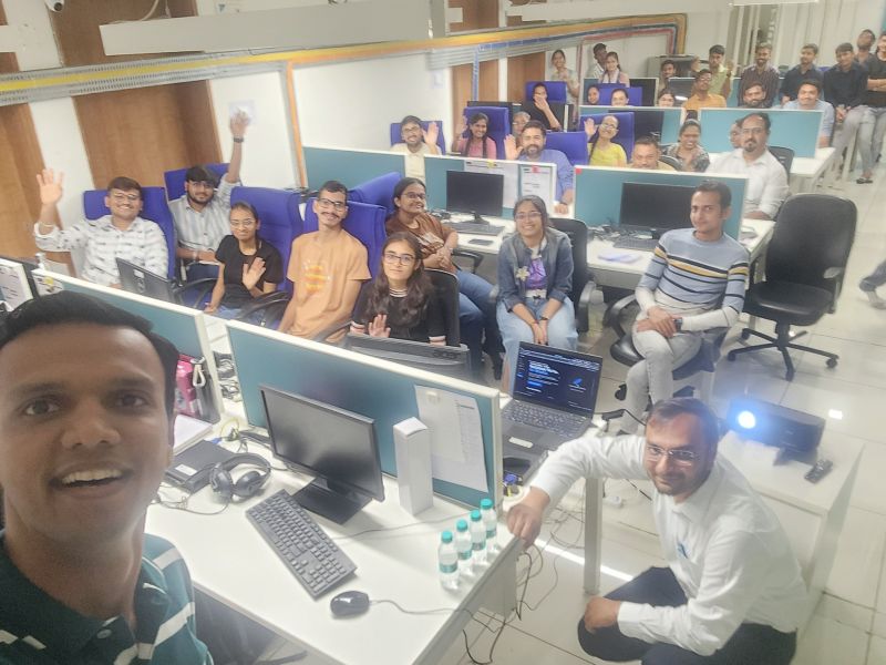 A man takes a group selfie with smiling colleagues waving from their desks in an open-plan office.