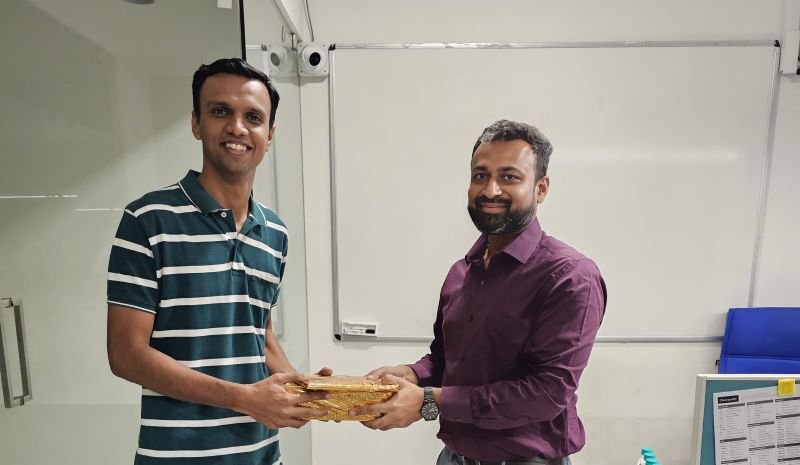 Two smiling men hold a wooden gift box together in an office in front of a whiteboard.