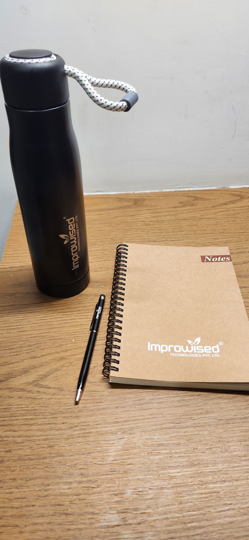 Black Improwised-branded water bottle, pen and spiral Notes notebook on a wooden table.