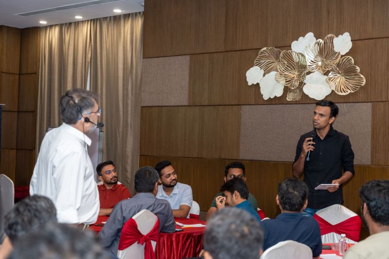A man holding a microphone speaks to seated attendees at a session in a wood-panelled room.