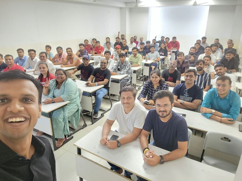 A smiling man takes a selfie with a large group of people seated at desks in a classroom-style room.