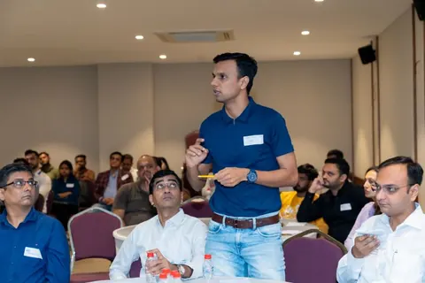 A man stands holding a pencil among seated attendees at a conference, appearing to ask a question.