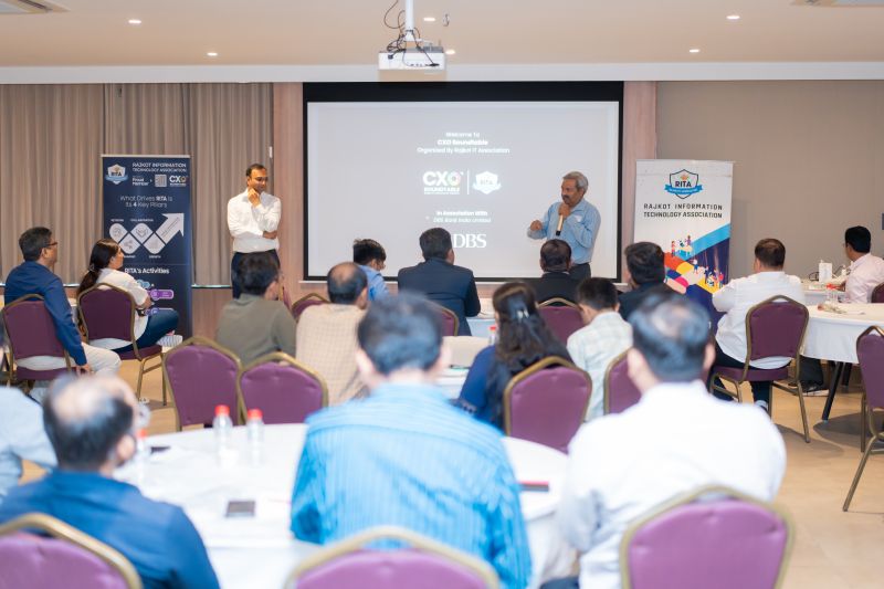 A man speaks into a microphone at a CXO roundtable event with association banners and a projection screen.