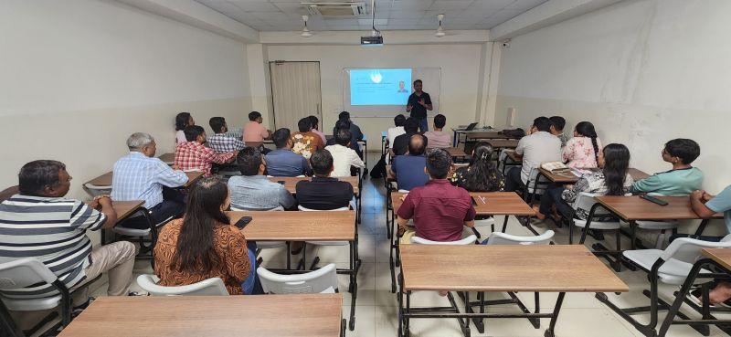 Speaker presenting beside a projector screen to an audience seated at classroom desks.
