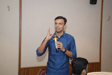 Man in a blue shirt speaking into a microphone and gesturing during an indoor session.