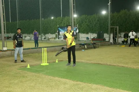 A batsman in a yellow and black jersey waits at the crease with a bat during a night box-cricket match on artificial turf.