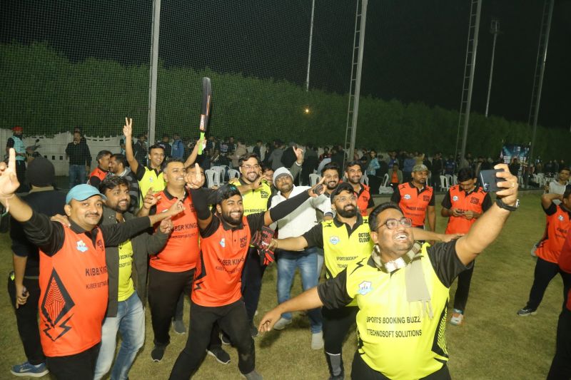 Men in orange and yellow team jerseys cheer with raised arms while one takes a selfie on a floodlit cricket ground at night.