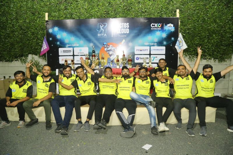 A cricket team in yellow and black jerseys sits in a row with trophies in front of an OCTOS CLOUD sponsor backdrop at night.