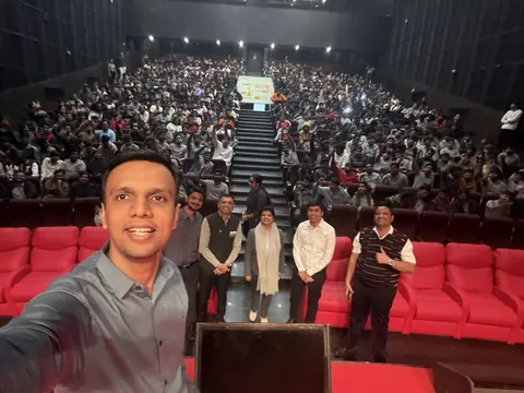 A man takes a selfie in front of a small group and a packed university auditorium with rows of red seats.